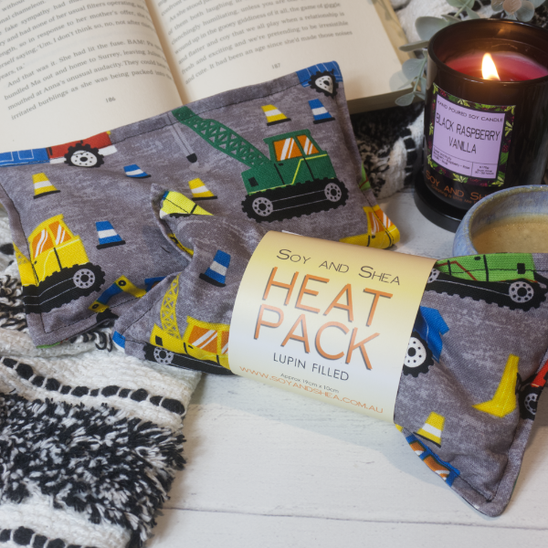 Two Mini Heat packs are stacked upon each other, one with a belly band label reading HEAT PACK LUPIN FIlled. These are resting on a black and white blanket with open book, candle and coffee cup evoking a cosy feel. The Heat packs are a dark grey mottled fabric with bright coloured contruction vehichles and traffic cones. Each pack shows a slight variation in pattern placement
