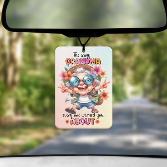 A mock up image showing a view through a car windscreen with a long tree lined road. From the rearview mirror hands an air freshener with an image of a grandma dressed as a hippie giving peace signs with the words THE CRAZY GRANDMA EVERYONE WARNED YOU ABOUT