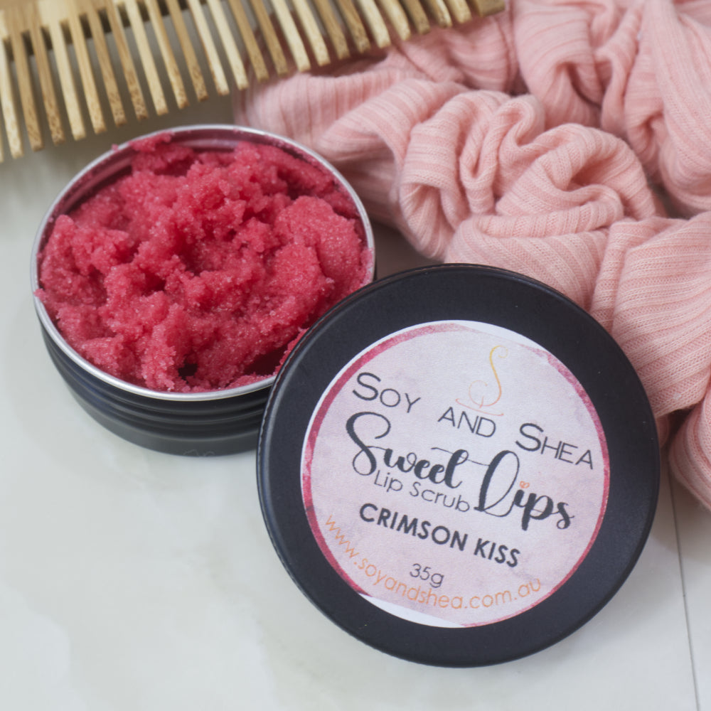 A black tin of Lip Scrub sits on a marble countertop next to a scrunchie and wooden comb. The lid has been removed and rests on the side of the tin revealing a red scrub