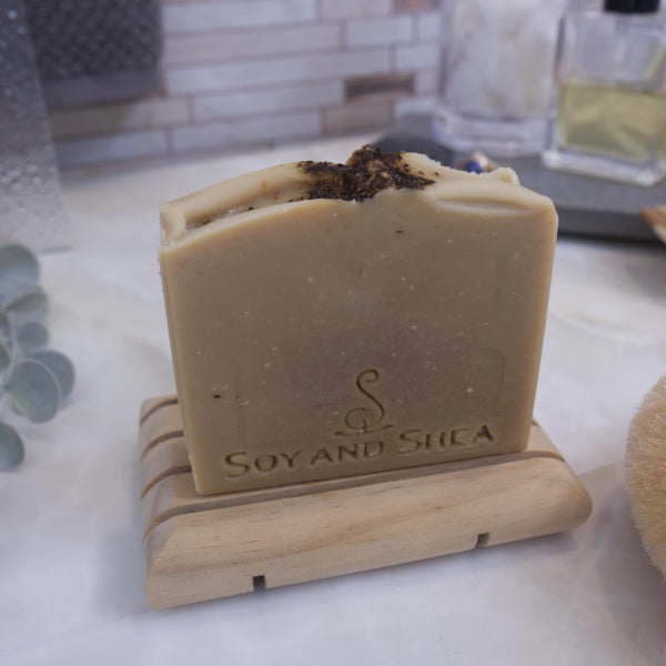 A pale khaki green sap bars stamped with SOY AND SHEA, sits on a wooden rack with a faded bathroom background. Tea leaves are sprinkled on the top of the soap