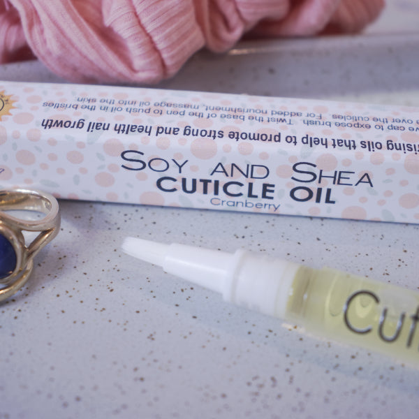 A close up image of the Cuticle oil pen showing the white brush tip. In the background is the box packaging with text CUTICLE OIL CRANBERRY and faded images of cranberries