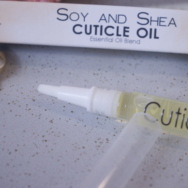 A close up image of the Cuticle oil pen showing the white brush tip. In the background is the box packaging with text CUTICLE OIL ESSENTIAL OIL BLEND