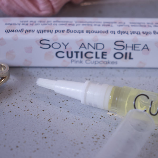 A close up image of the Cuticle oil pen showing the white brush tip. In the background is the box packaging with text CUTICLE OIL PINK CUPCAKES and faded images of cupcakes with pink base and white icing