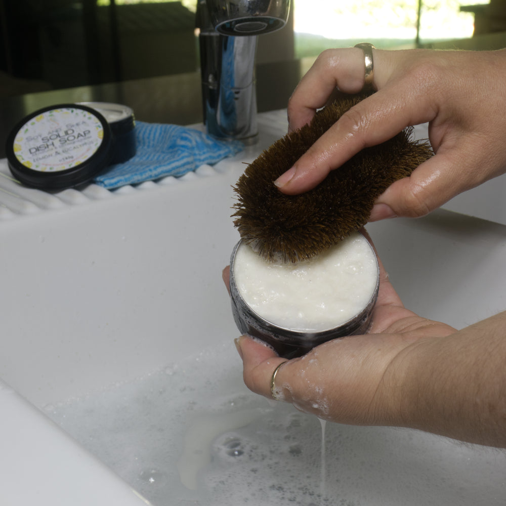 Person scrubbing a container of soap with a brush over a sink.