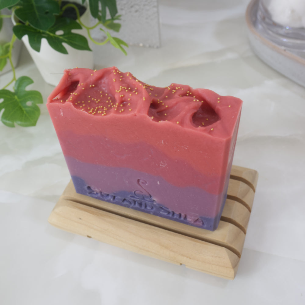 A bar of soaps stands on a wooden soap rack on a bathroom countertop along with a vine-like plant and stone tray. The soap is pictured from above showing a red top with gold cellulous beads. The soap has four jaggered layers of colour starting with dark then light purple. Followed by light then bright red.