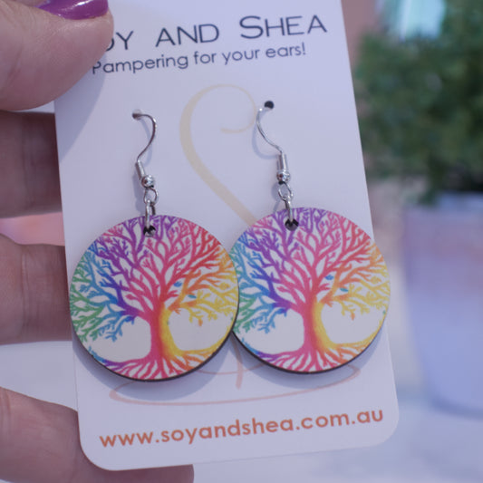 A hand holds a white card branded with SOY AND SHEA with a pair of earrings hanging on hooks. The earrings are round with a rainbow coloured "Tree of Life" symbol against a white background