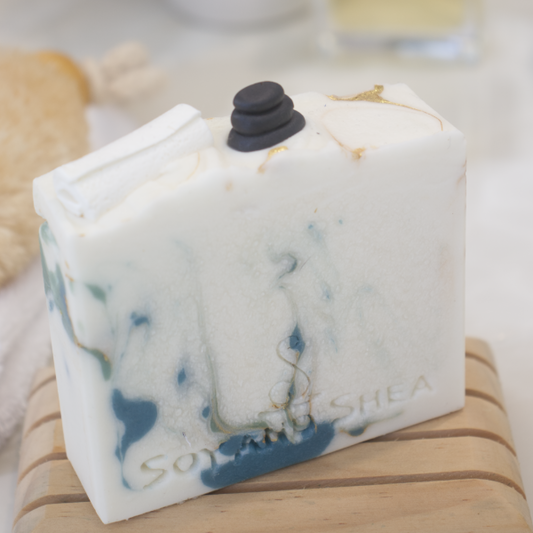 A close up photo of the Elysian Soap bar with a blurred background. The soap is off white with dark green and gold swirls. The top of the soap features soap embellishments of a stack of massage stones and a rolled up white towel.