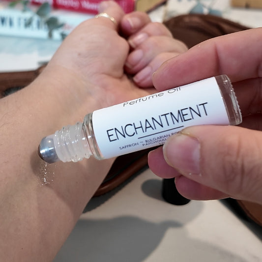 A person is applying a roller bottle of perfume oil labelled ENCHANTMENT to the other wrist.