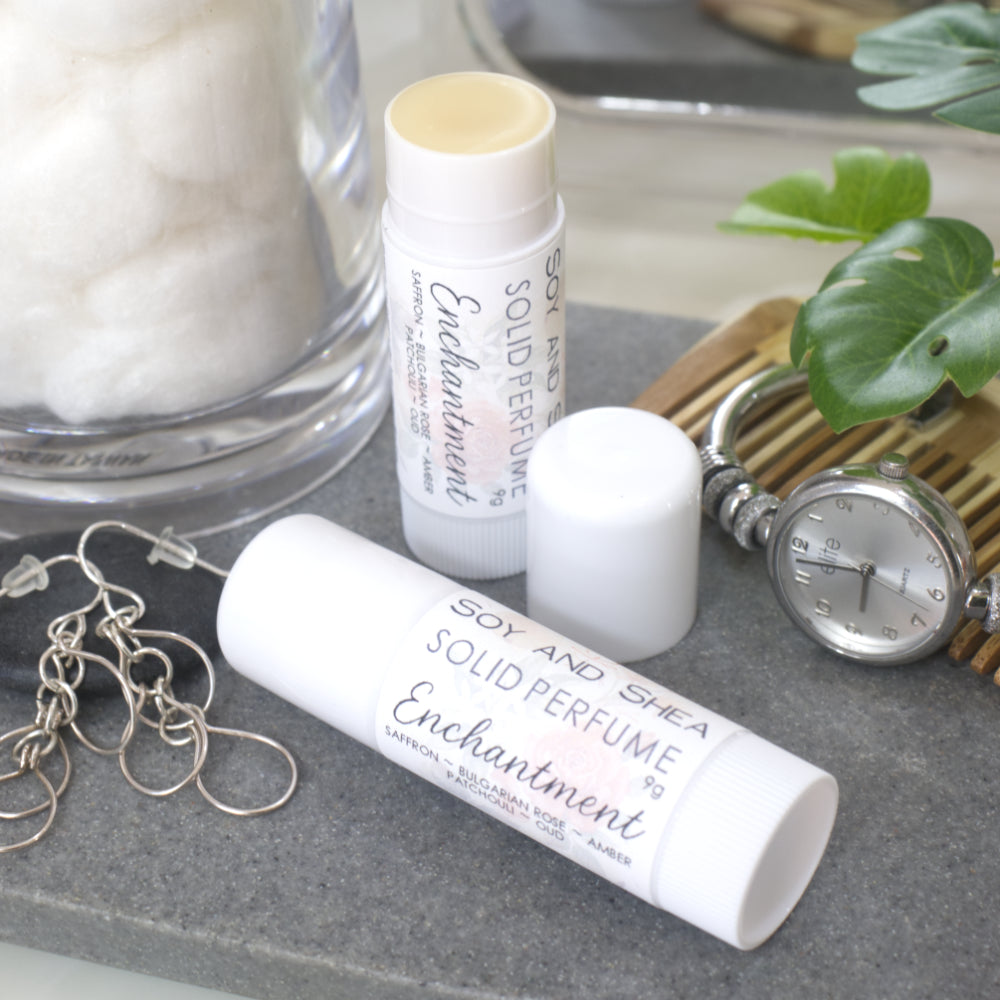 Two white tubes of Solid Perfume are displayed on a grey stone tray along with silver earrings, watch and greenery. One tube lays flat displaying the fragrance notes while another tube stands upright behind it with the lid removed and to the right revealing a beige balm