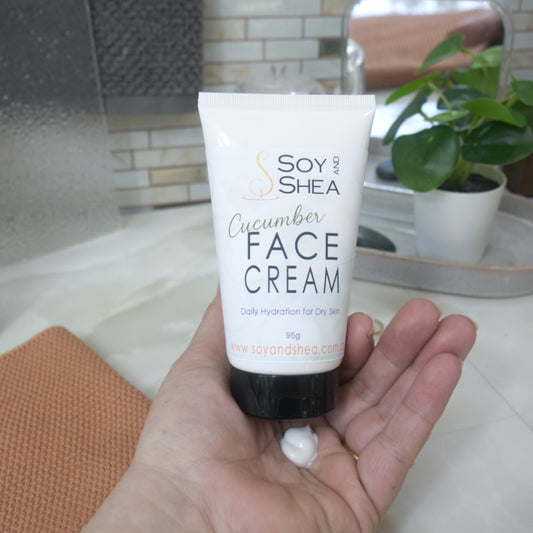 A hand holding a white squeeze tube of Soy and Shea Cucumber Face Cream, with a small dollop of white cream resting on the palm. The background shows a softly blurred bathroom setting with a sink, tiled wall, and a potted plant.