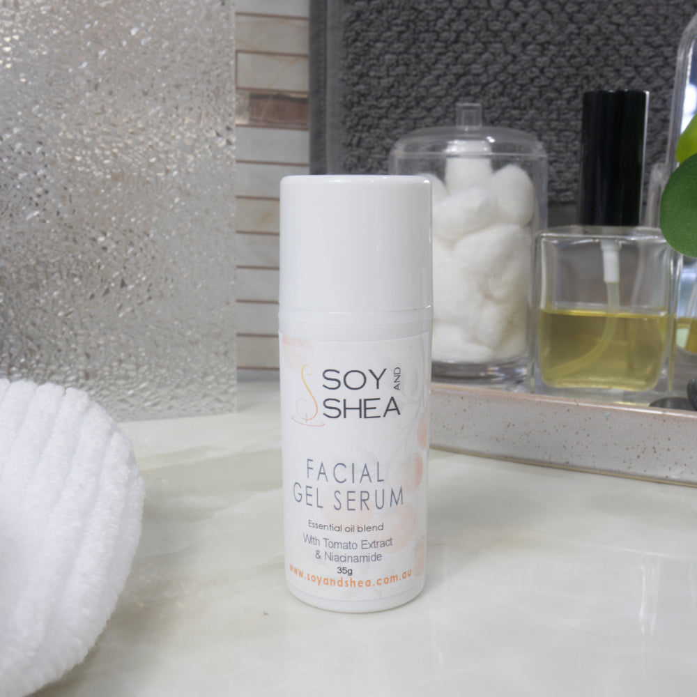 A white cylindrical bottle of Soy Shea Facial Gel Serum stands on a light countertop, with cotton pads and a small bottle of oil in the background.