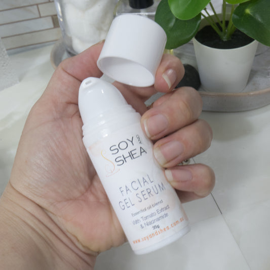 A hand holding a white bottle of Soy & Shea Facial Gel Serum with its cap off, set against a bathroom counter background with a small green plant. The bottle it about the size of the palm of the hand and has a dispensing pump under the cap