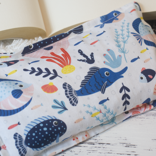 A close up image of the Mini Fish Heat pack highlighting the fabric design which shows illustrations of a variety of reef fish in shades of blue along with pieces of coral on a white background.