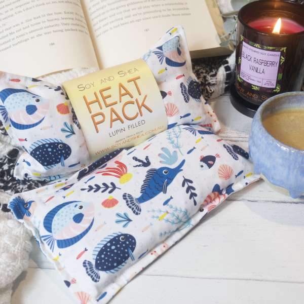 Two Mini Heat Packs sit on a wooden bench resting against a black and white blanket with an open book. A burning candle and cup of coffee complete the cosy scene. The Heat packs are rectangular in shape with a white background covered with illustrations of a variety of reef fish and coral in shades of blue. One Heat Pack shows the packaging which is a simple belly band with the text HEAT PACK LUPIN FILLED