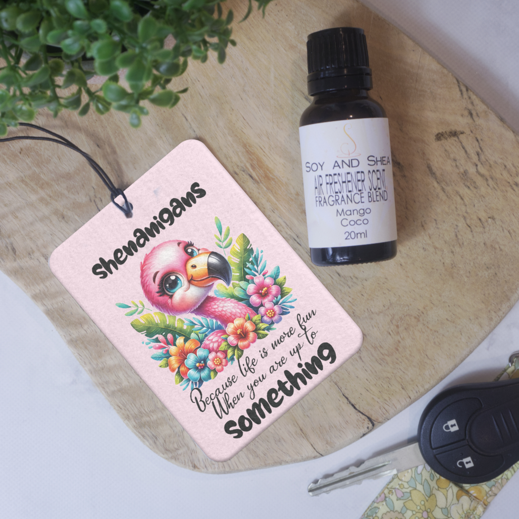 A car air freshener lies on a wood surface with an illustration of a cartoon flamingo surrounded by flowers and the words SHENANIGANS BECAUSE LIFE IS MORE FUN WHEN YOU ARE UP TO SOMETHING. Next to the air freshener is a bottle of scent