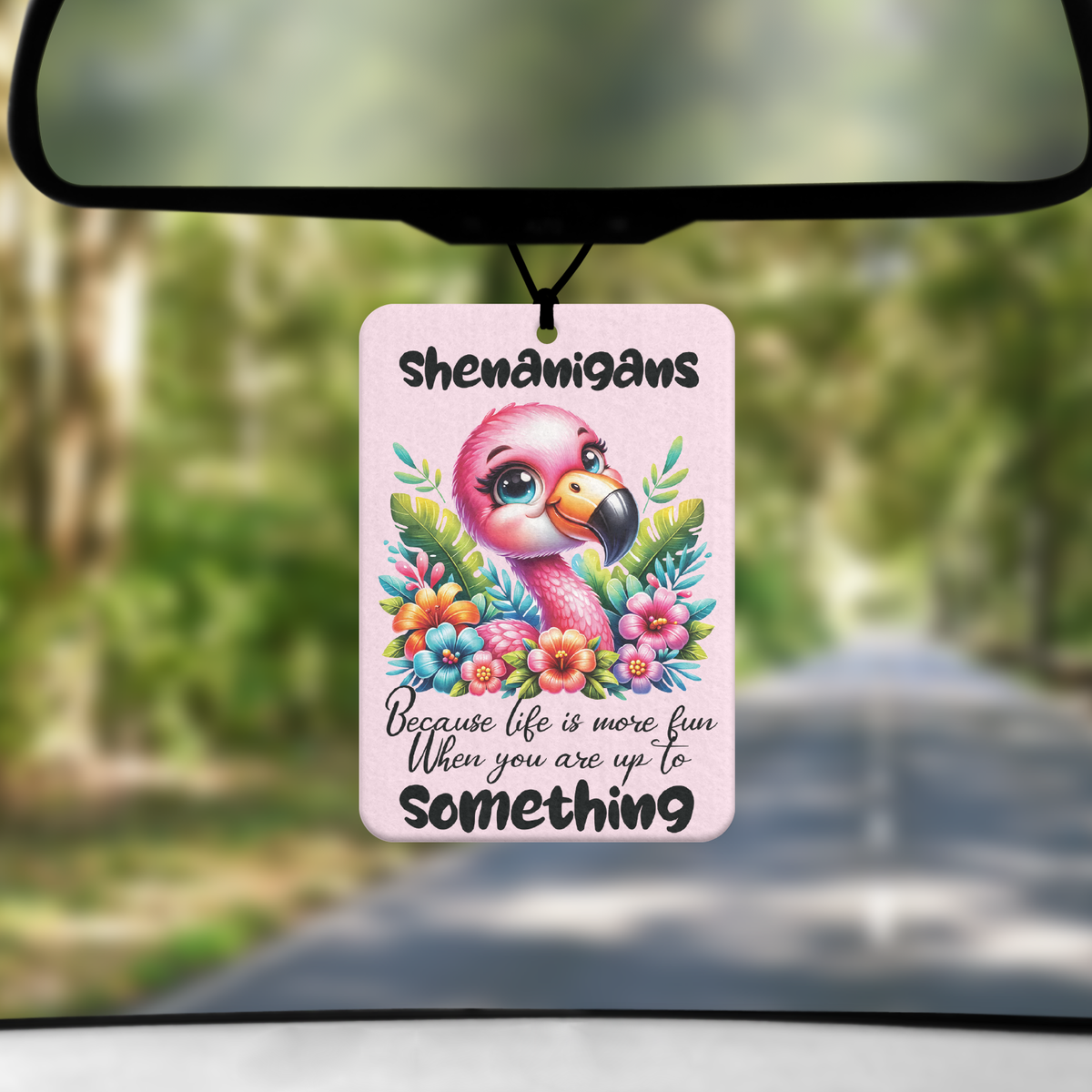 A mock up image showing a view through a car windscreen with a long tree lined road. From the rearview mirror hangs an air freshener with an image of a pink flamigo surrounded by flowers and the text SHENANIGANS BECAUSE LIFE IS MORE FUN WHEN YOU ARE UP TO SOMETHING