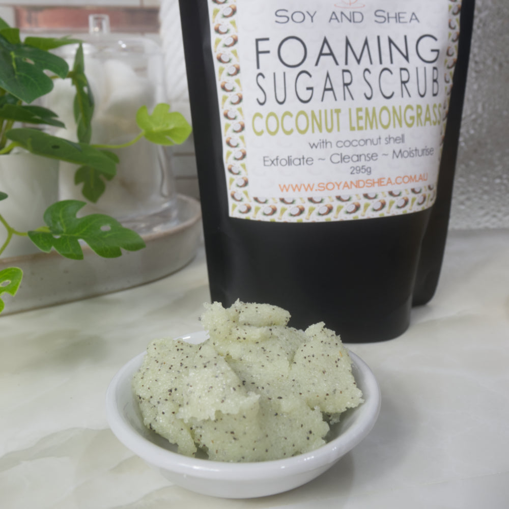 A close up image of a small white bowl filled with a pale green body scrub that features brown flecks of coconut shell. Behind is a slightly blurred image of a black pouch bag with a product label identifying this has a Foaming Sugar Scrub scented with Coconut Lemongrass.