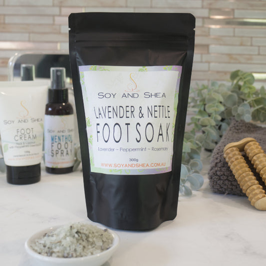 A black zip lock pouch of Soy and Shea Lavender and Nettle Foot Soak sits on a bathroom counter with Foot Cream and a bottle of Foot Spray in the background against a decorative tile wall and greenery. In the foreground is a small white dish filled with salt speckled with botanicals.