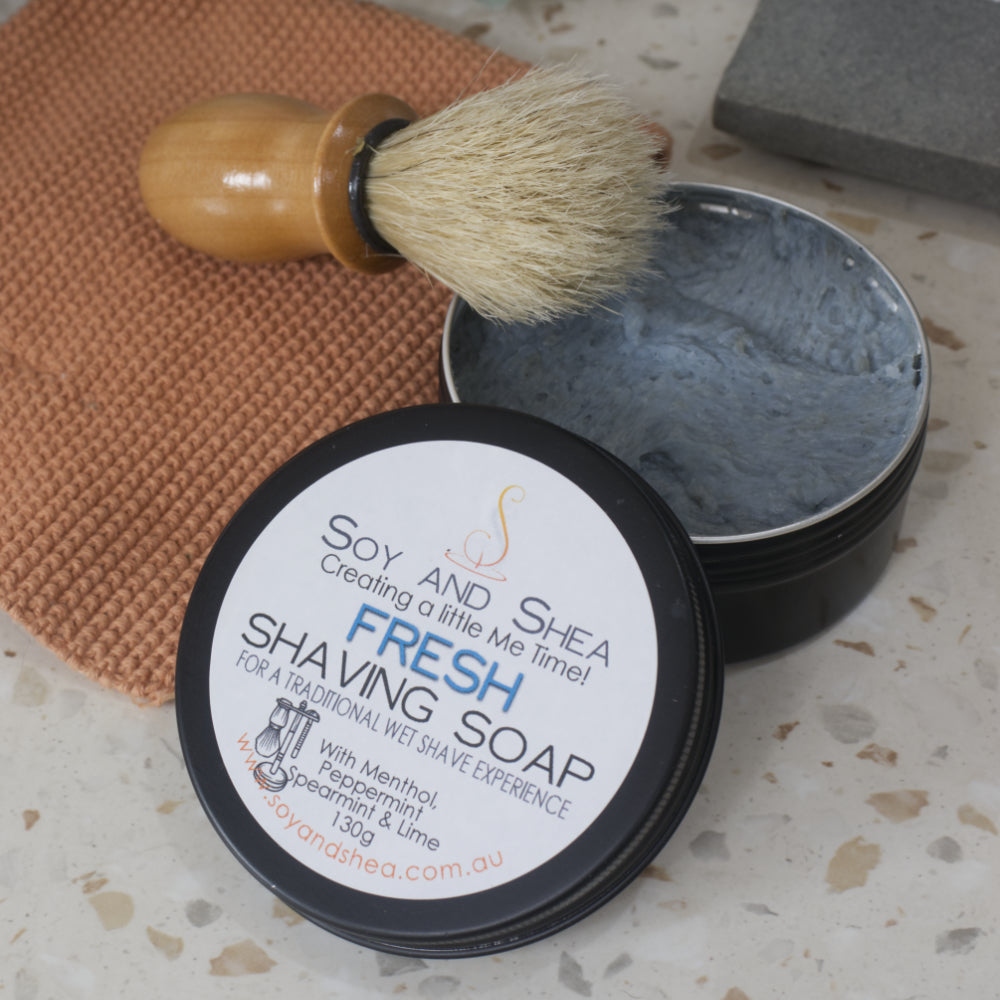 A black tin of fresh Shaving Soap sits on a bathroom counter top with the lid off and resting on the side. The Soap is a blue colour. Resting on the tin is a shaving brush