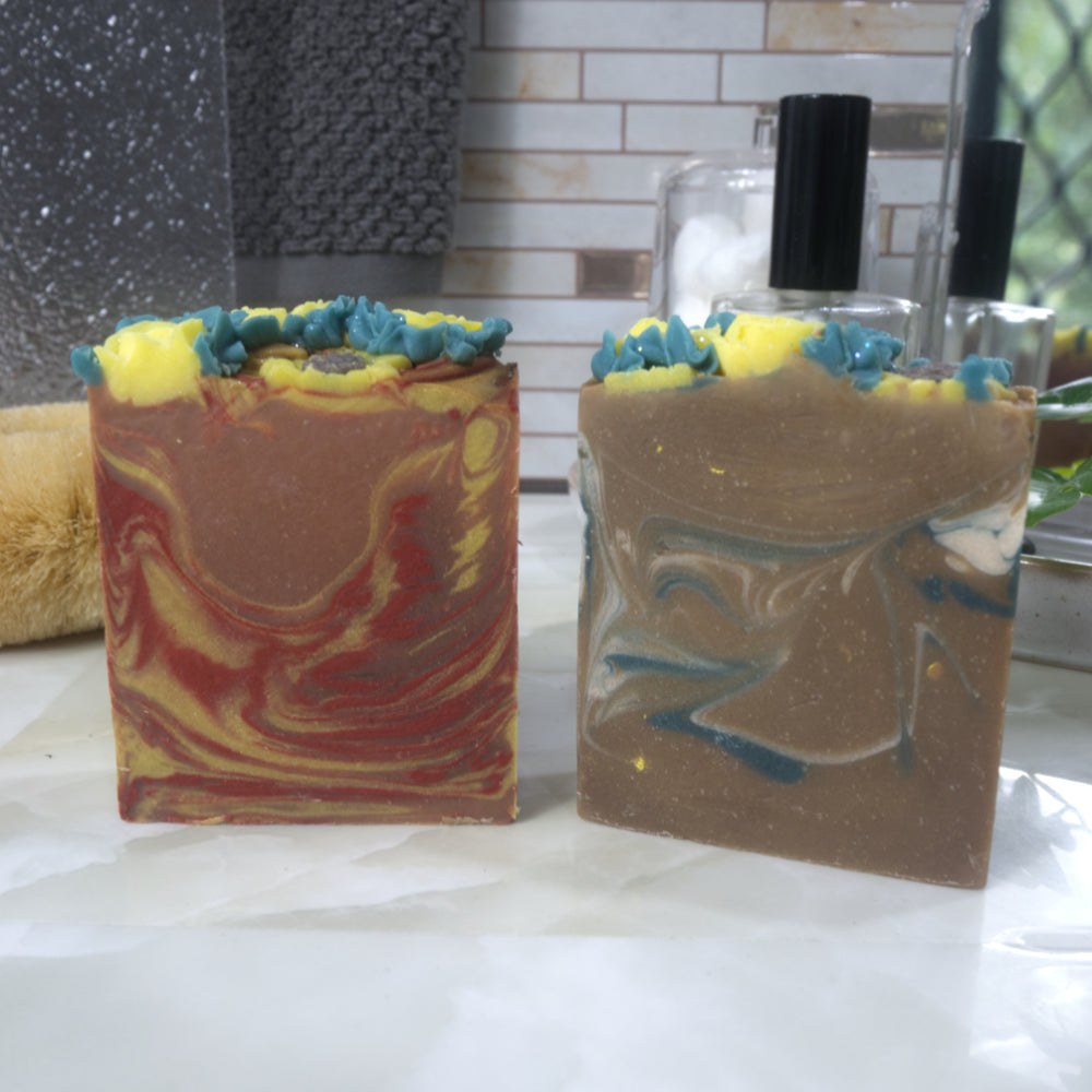 Two bars of soap stand upright, side-by-side on bathroom counter top. The soap to the left has swirls of orange, red and yellow, while the other has green and white swirls through a rich gold base. The top has piped yellow flowers and green leaves