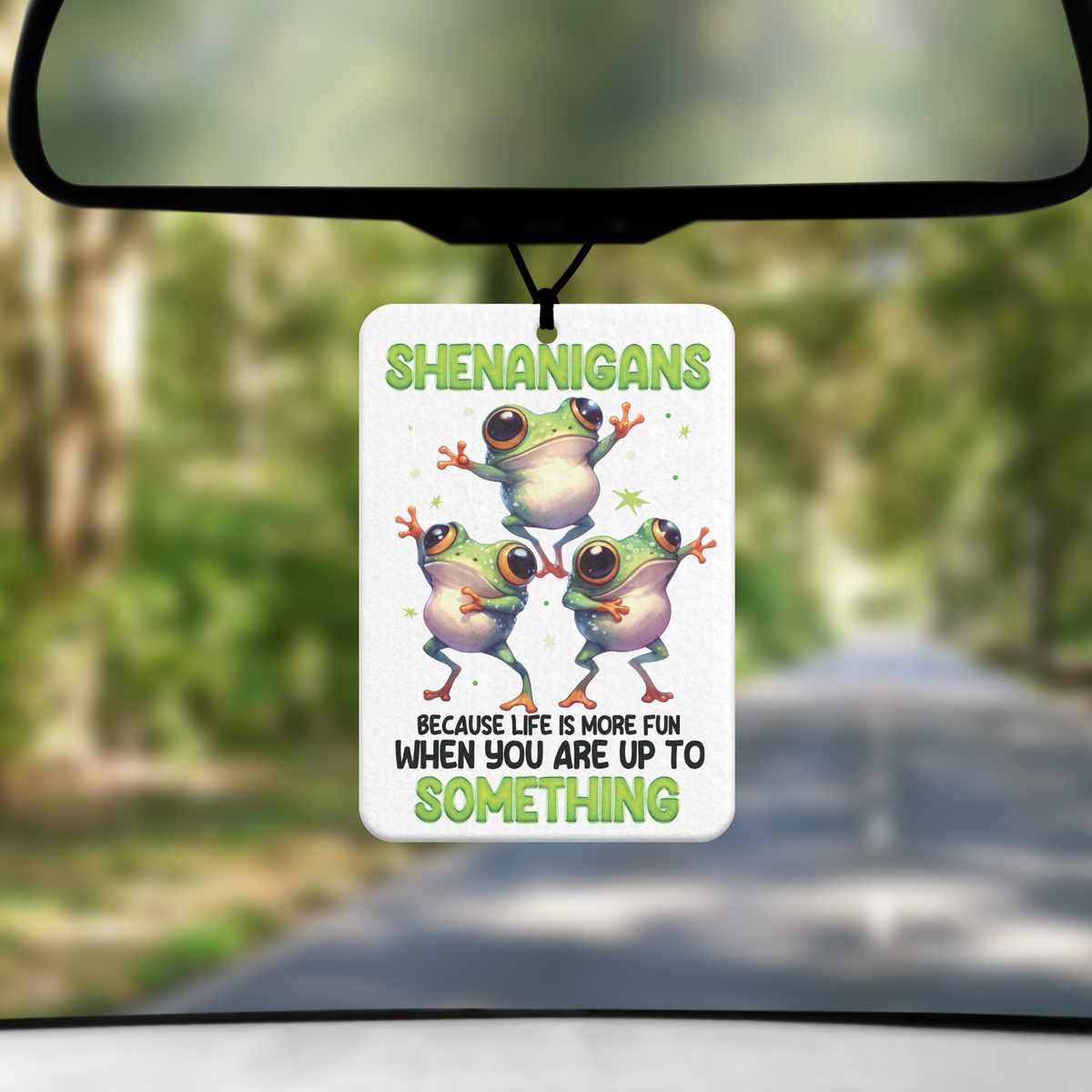 A mock up image showing a view through a car windscreen with a long tree lined road. From the rearview mirror hangs an air freshener with an image of three frogs dancing with the word Sheananigans across the top in green. Along the bottom it says "because life is more fun when you are up to SOMETHING"