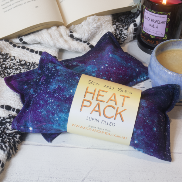 Two Mini Heat packs are stacked upon each other, one with a belly band label reading HEAT PACK LUPIN FIlled. These are resting on a black and white blanket with open book, candle and coffee cup evoking a cosy feel. The Heat packs have a mottled blue to purple design with splashes of white dots to represent stars