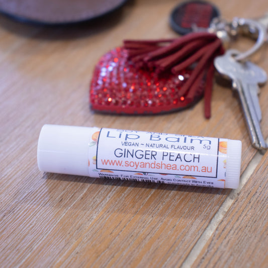A white tube of lip balm lies on a wooden surface next to a key on a red heart shaped keyring. The lip balm label is white with watercolour images of peaches.