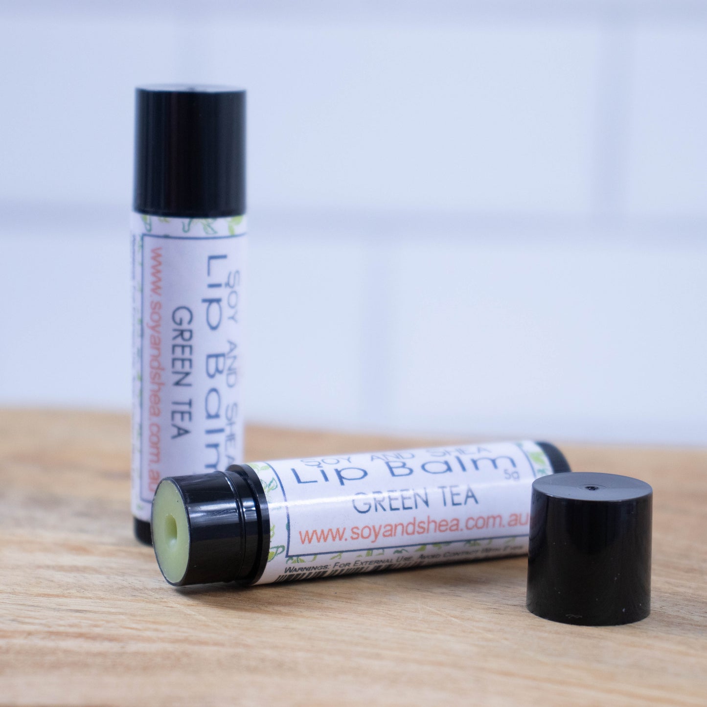 Two black tubes of lip balm are displayed on a wooden surface with blurred tiled background. One tube stands on its end while the other lays flat in front with the cap removed revealing a light green balm. The label is white with a random pattern of green leaves.