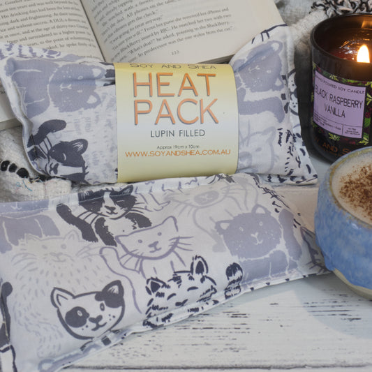 Two Grey Cats Mini Heat Pack by Soy and Shea rests near a ceramic cup, a burning candle, and an open book—creating an inviting cosy scene. The Mini Heat packs feature a white background with sketches of a variety of cats in grey. Each pack shows variations in the design including one horizontal and the other vertical.