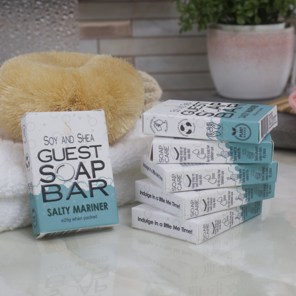 To the left, a box of Salty Mariner Guest Soap bar rests against a while towel with body brush. The right is a stack of 5 other boxes.
