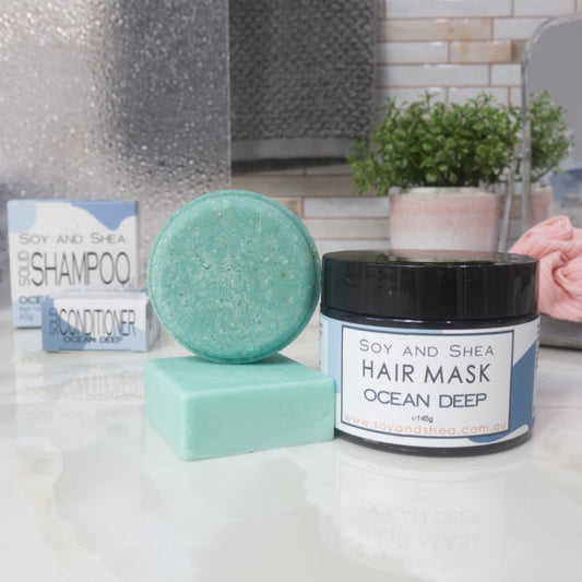 Hair care products including a bar of shampoo, a bar of conditioner, a jar of hair mask, on a bathroom counter. All products are in shades of blue-green.