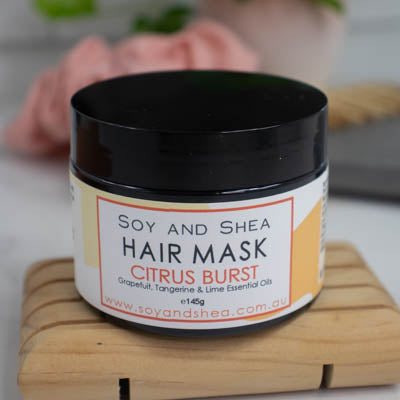A black jar with lid on sits on a wooden surface against a blurred background with scrunchie and comb. The jar features a white label with a yellow abstract design with text HAIR MASK CITRUS BURST.
