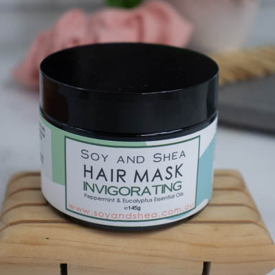 A black jar with lid on sits on a wooden surface against a blurred background with scrunchie and comb. The jar features a white label with a green abstract design with text HAIR MASK INVIGORATING.