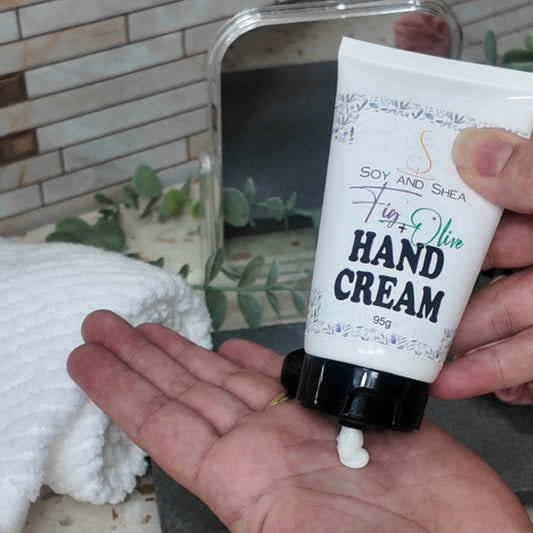 Hand holding a white tube of Fig & Olive Hand cream with a small amount squeezed out onto the palm of the other hand, against a bathroom counter background.
