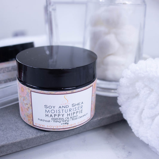 A black jar of with a floral labels sits on a grey stone tray surrounded by a white towel, cotton balls and a mirror reflecting the back of the jar