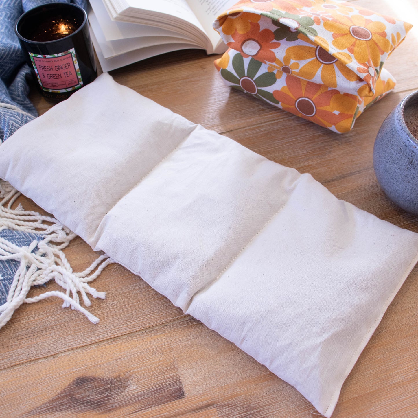 The insert of a Heat Pack lays on a wooden surface next to a burning candle, open book and filled coffee cup. To the right is a folded heat pack. The insert is a long rectangular shape made of a cream fabric. It has two lines of stitching going from long side to long side dividing the insert into 3 equal parts.