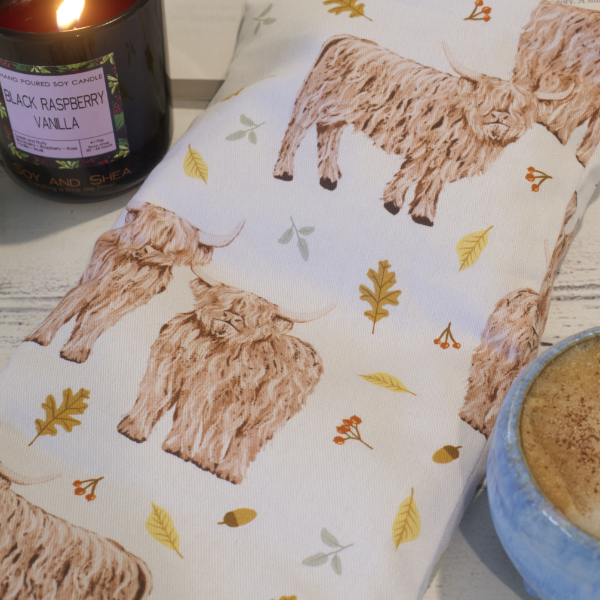 A close up image of a Highland Cow Heat Pack next to a burning candle and cup of coffee. The heat pack has a illustrations of Hightland Cows on a white background surrounded by Autumn folliage