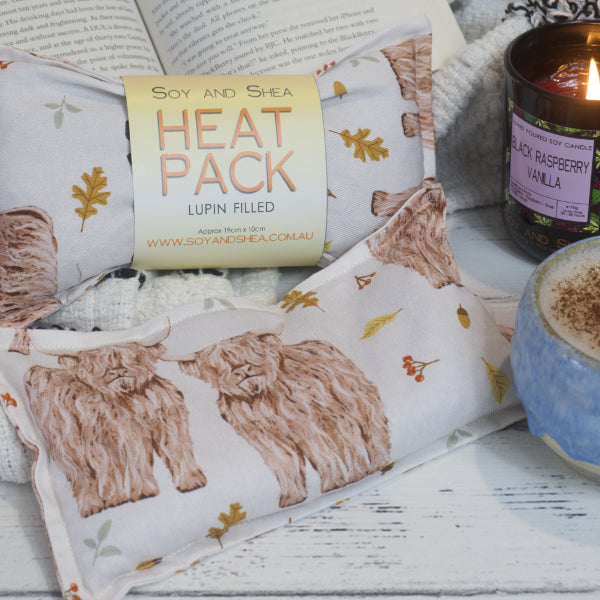 The Soy and Shea Highland Cows Mini Heat Pack sits next to a burning black raspberry and vanilla soy candle, an open book, and coffee on a white wooden surface. There are two heat packs, one with a label, showing a white rectangular pack with Highland Cows and autumn folliage including acorns.