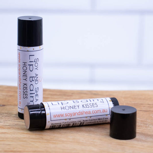 Two black tubes of lip balm are displayed on a wooden surface with blurred tiled background. One tube stands on its end while the other lays flat in front with the cap removed revealing a light brown balm. The label is white with a random pattern of bees in flight