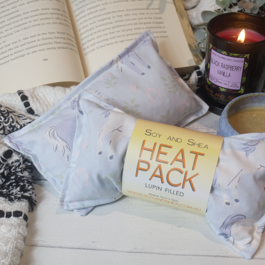 Two Mini Heat Packs rest on a wooden table top resting against an open book surround by a black and white blanket, burning candle and filled coffee cup. The heat packs are a light blue colours with illustrations of white and blue horses. The Heat Pack towards the front has a belly band label reading HEAT PACK LUPIN FILLED