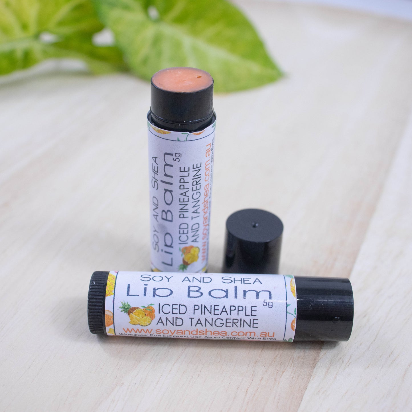 Two black tubes of lip balm displayed on a wooden surface with greenery blurred in the background. One tube lies flat across the surface displaying the flavour while the other tube stands upright behind it with the cap removed and placed to the side revealing an orange balm.