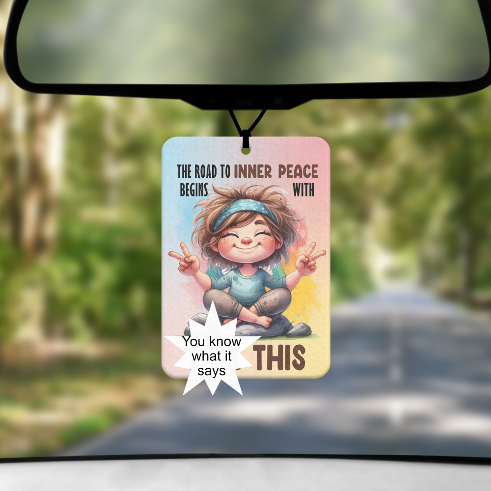 A mock up image showing a view through a car windscreen with a long tree lined road. From the rearview mirror hangs an air freshener with an image of a lady dressed like a hippie with the words "the road to inner peace begins with ... this. The one word has a star over it