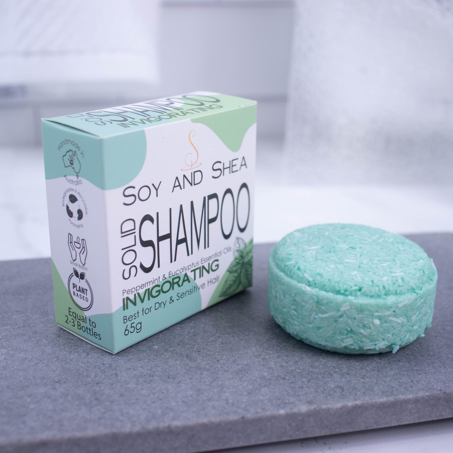 A round mint green Shampoo bar and its cardboard box which is white with an abstract green design are displayed on a grey stone tray with a blurred bathroom background.