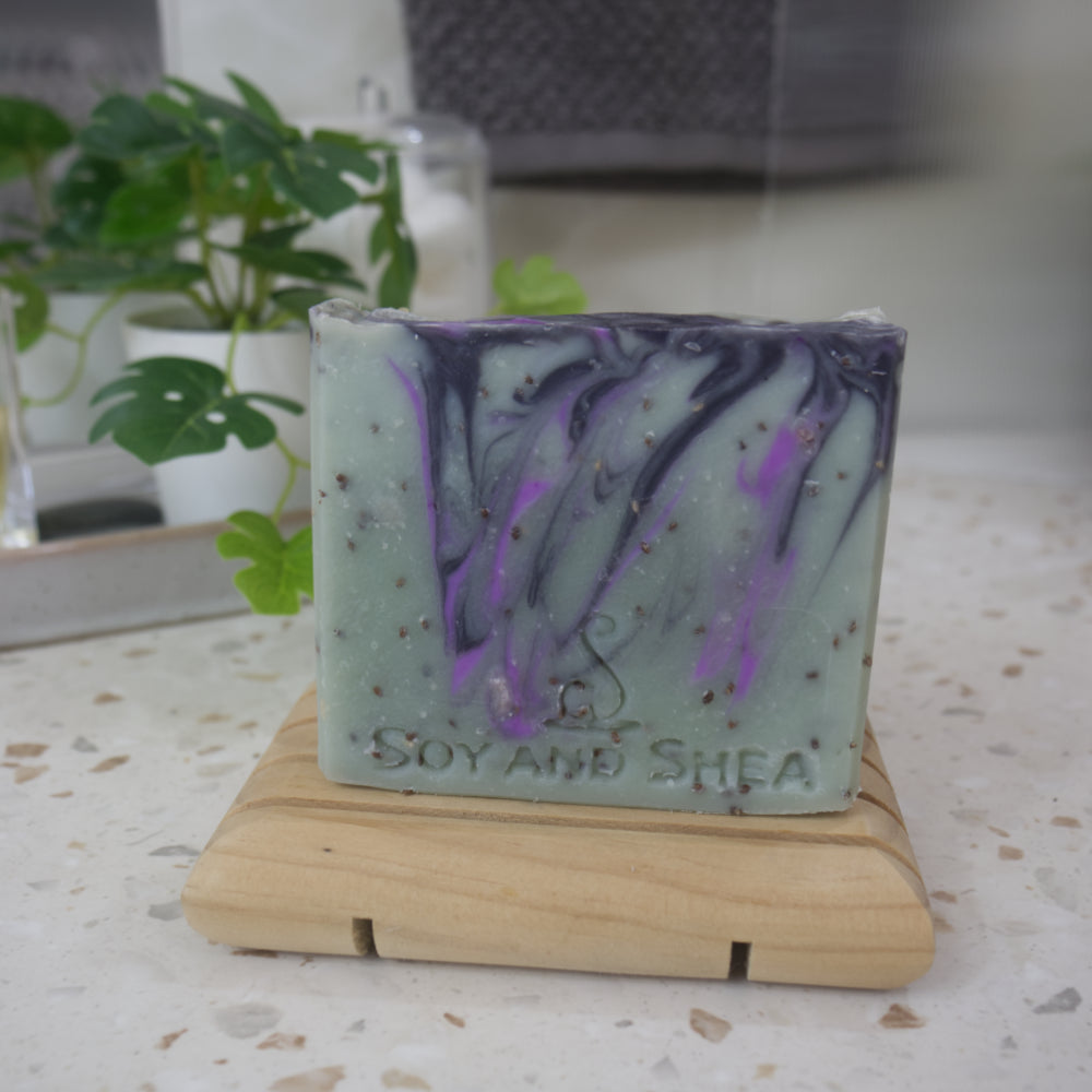 A bar of soap stands on a wooden soap dish next to a plant on a bathroom countertop. The soap has a forrest green base colour with brown flecks along with light and dark purple swirls of colour.