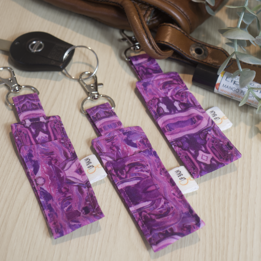 A selection of three clip balms showing variations in fabric design, lie on a wooden side with the middle Clip Balm attached to a car key. A lip balm lays near-by along with some greenery. The Clip balms are various shades of bright to dark purple in a swirl design