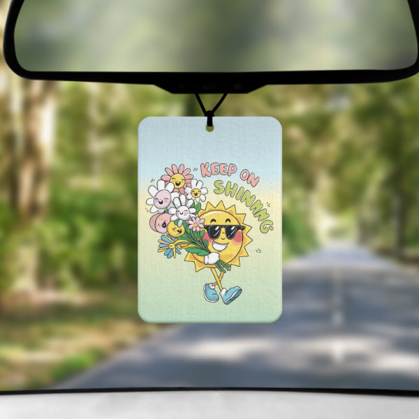 A mock up image showing a view through a car windscreen with a long tree lined road. From the rearview mirror hangs an air freshener that has an ombre background of blue at the top green towards the bottom. It has a cartoon image of a smiling sun walking along with a bunch of flowers all with smiling faces next to text reading KEEP ON SHINING
