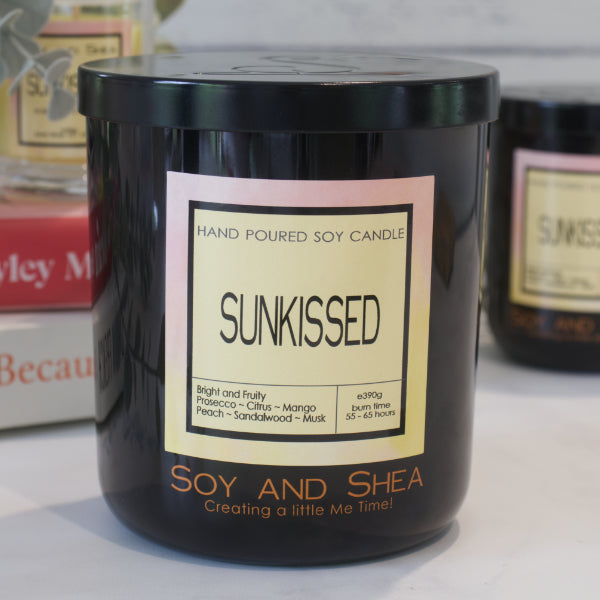 A black candle jar with a lid embossed with the Soy and Shea Logomark sits on a white counter with a label reading SUNKISSED and 390g, sits on white counter with a stack of candles in the background