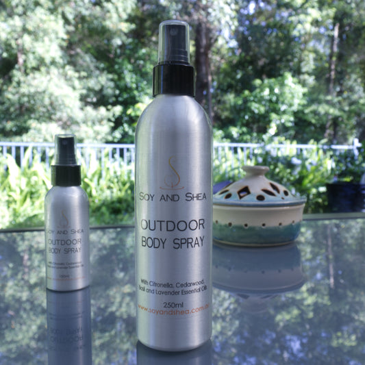 Two metal bottles of "Outdoor Body Spray" by Soy and Shea, stand on a glass table top with a lush green garden in the background. A large bottle sits in the front of the images with an atomiser style lid and reads 250ml, while a smaller bottle sits towards the back slightly blurred.