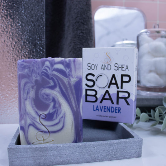 A close up look of the soap which has lavender, dark purple and white swirls. The soap is rectangular in shape and is taller than wide. It is sitting on a stone soap dish and behind it is the box the soap came in. The lower portion of the box has a bubble border coloured in lavender whereas the top is white with small grey bubbles. The box advises it is soap and the scent.