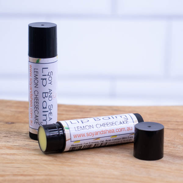 Two black tubes of lip balm are displayed on a wooden surface with blurred tiled background. One tube stands on its end while the other lays flat in front with the cap removed revealing a yello balm. The label is white with illustrations of slices of cheesecake